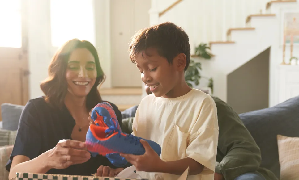Smiling boy holding a blue and red soccer shoe while a woman helps him with laces in a cozy living room.
