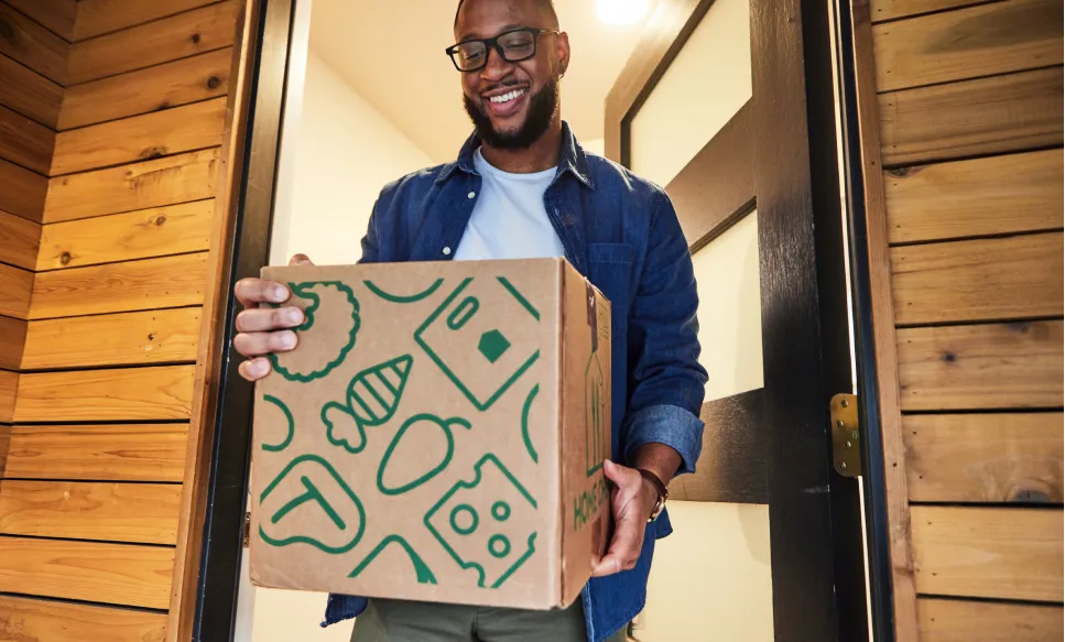 Smiling man wearing glasses and denim shirt holding a cardboard box with green food icons at a wooden doorway.