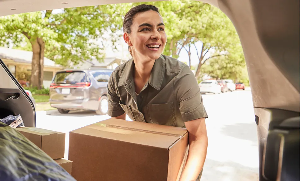 Smiling delivery woman holding a cardboard box by a vehicle in a sunny residential area.