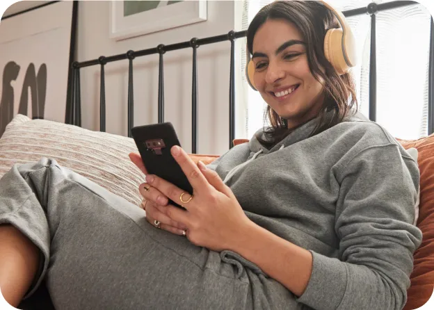 A woman sitting and smiling on a couch wearing headphone and using her phone.