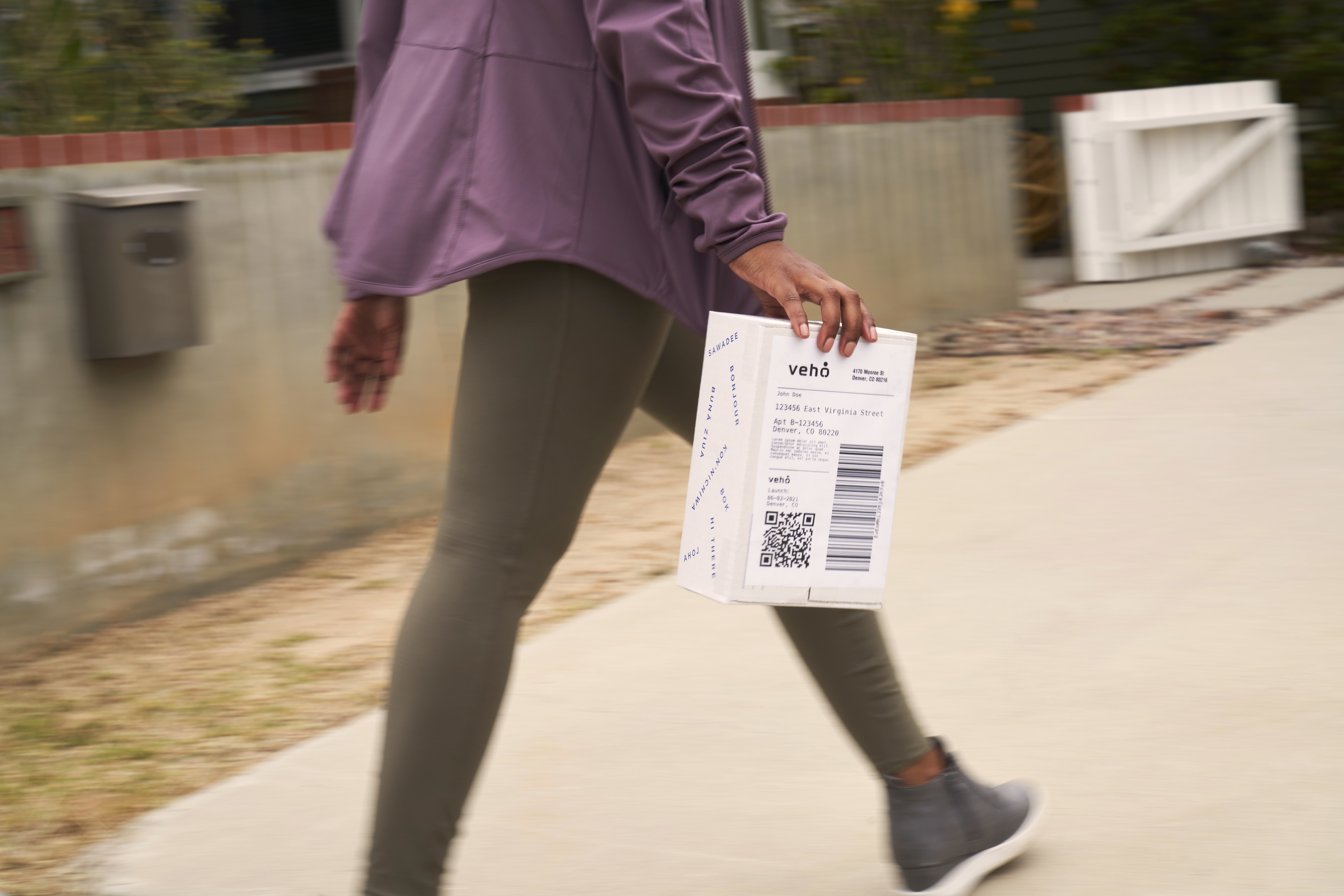 Person walking on sidewalk carrying a small white package labeled 'vehó' with barcode and QR code.