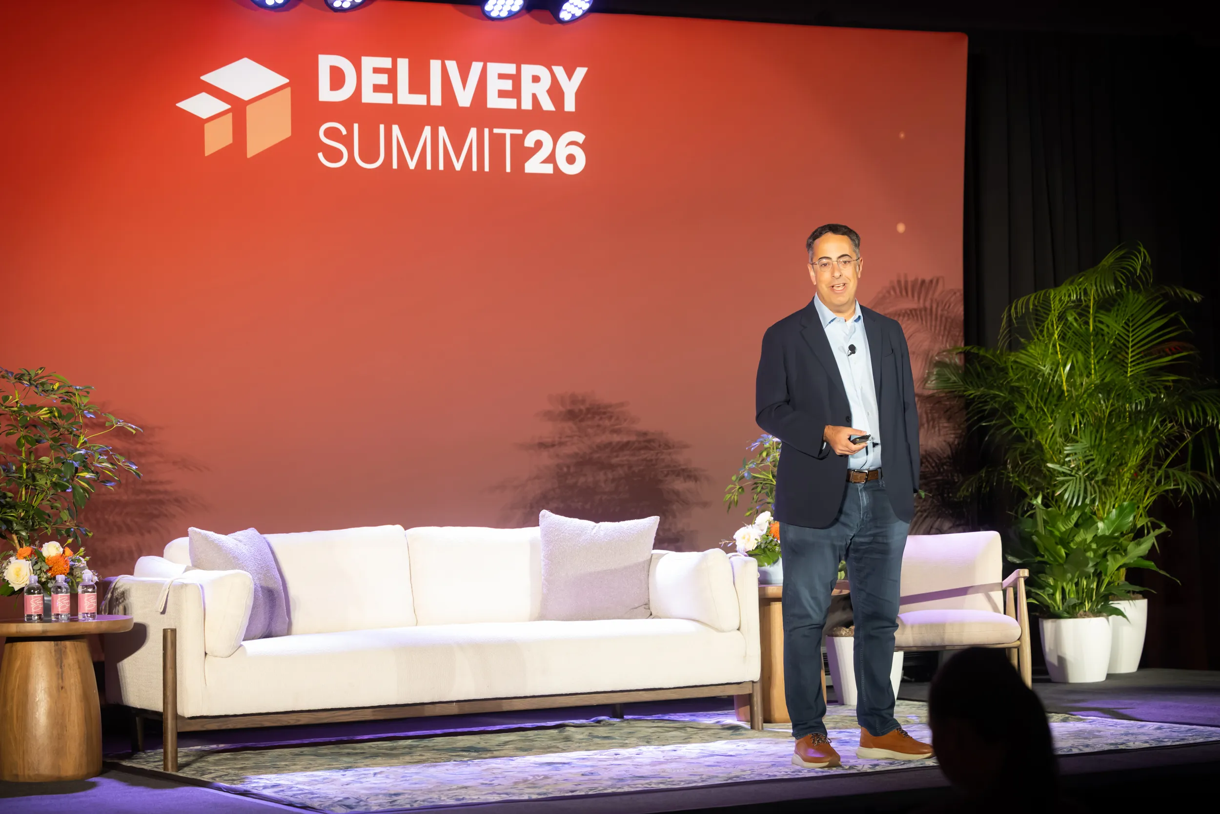Man in blazer and jeans speaking on stage at Delivery Summit 26 with sofa and plants in background.