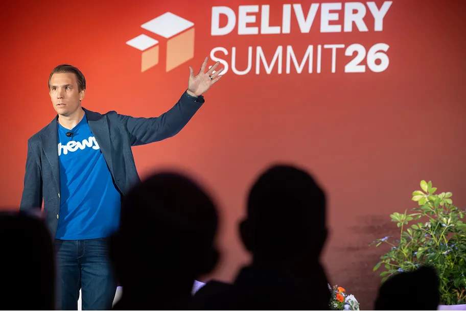Man in blue shirt and blazer speaking on stage at Delivery Summit 26 with raised hand gesture.