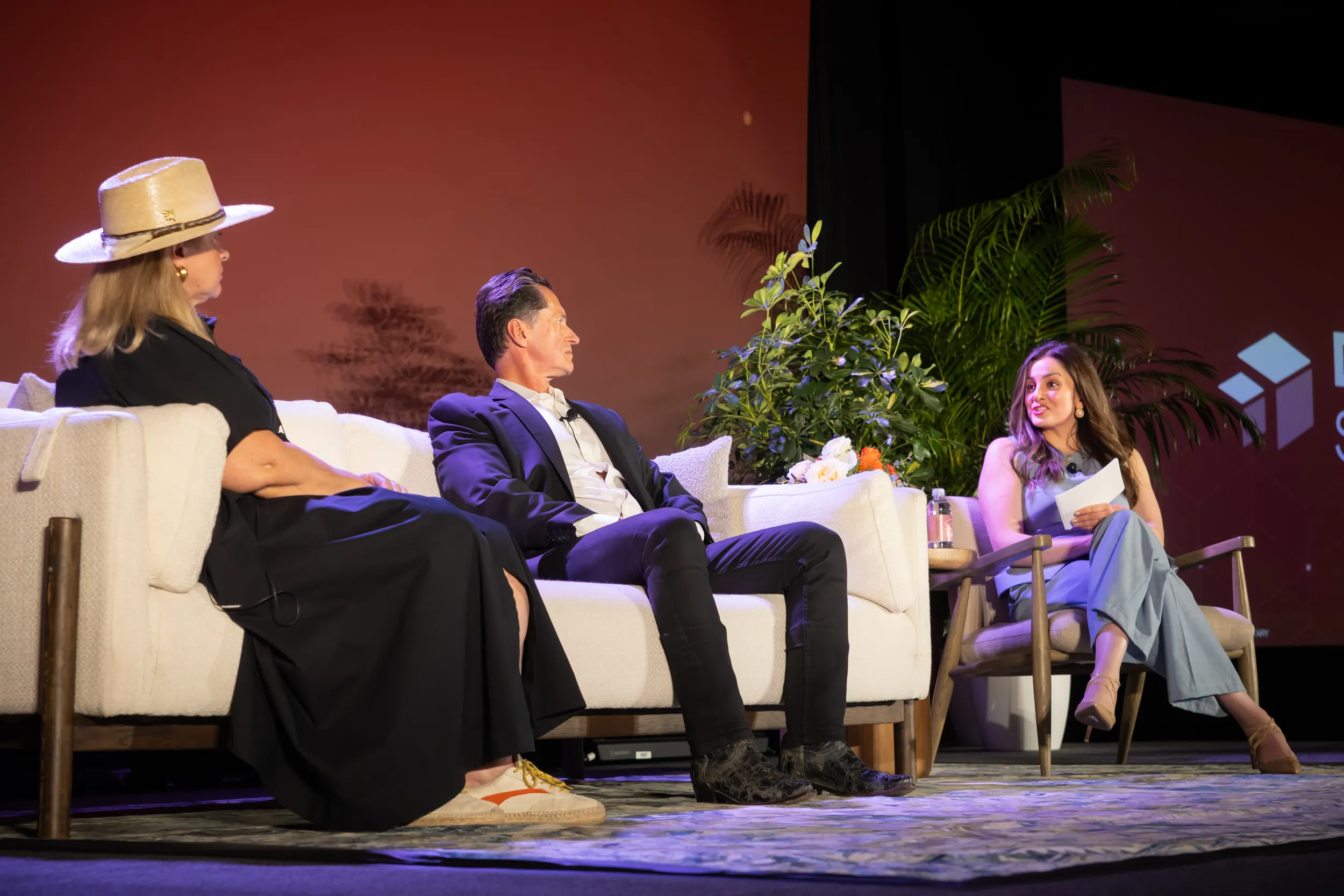 Three people seated on stage having a discussion, with two on a couch and one in a chair holding notes.