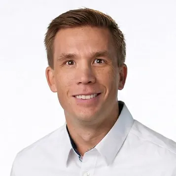 Smiling man with short brown hair wearing a white collared shirt against a white background.