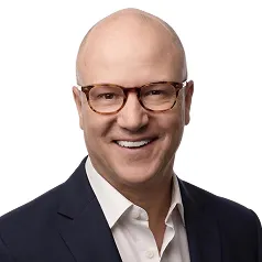 Smiling bald man wearing glasses, a white shirt, and a dark blazer against a white background.