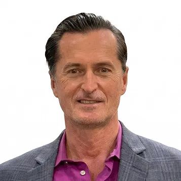 Middle-aged man with dark slicked-back hair wearing a gray blazer and a purple shirt.