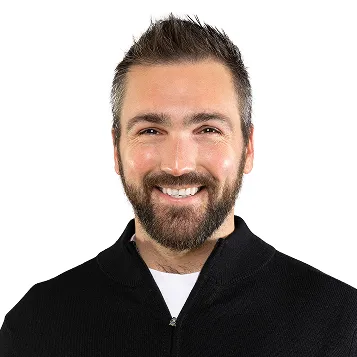 Smiling man with short brown hair, beard, and black zip-up sweater on white background.