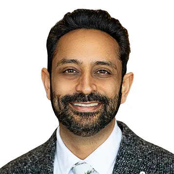 Smiling man with dark hair and beard wearing a gray suit jacket and white shirt with a tie.