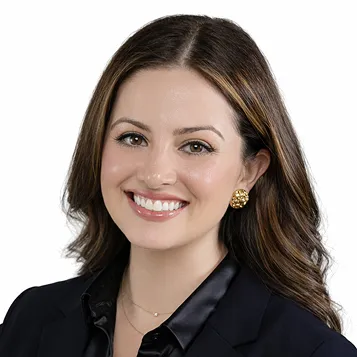 Smiling woman with long brown hair, wearing gold earrings and a black blazer on a white background.
